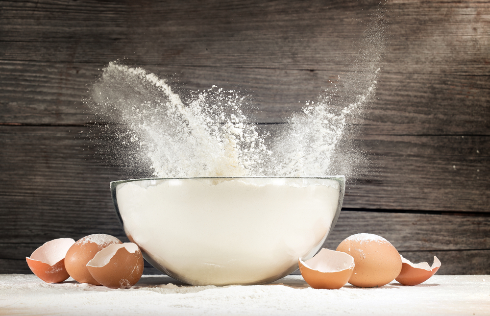 White,Flour,Splashing,Out,Of,A,Glass,Bowl,And,Eggshells